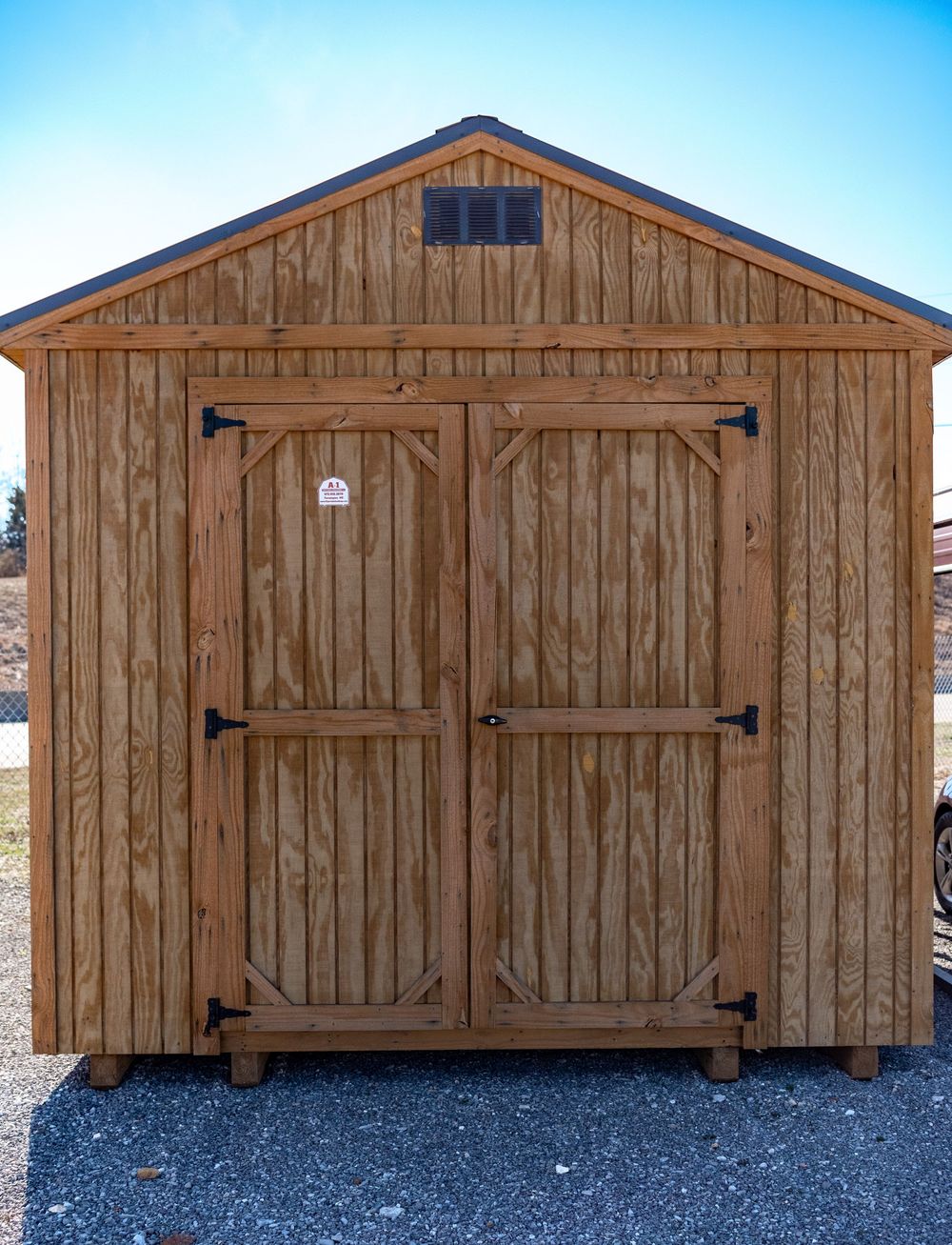 Wooden storage shed with double doors, black hardware, and a dark shingled roof on a gravel surface.