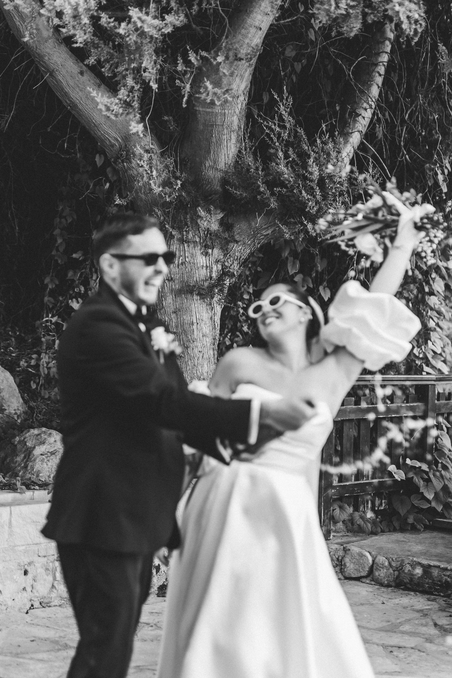 A couple in wedding attire dancing playfully in black and white under a tree.