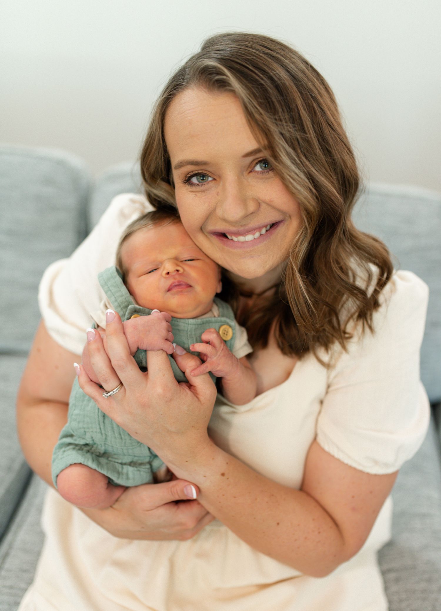 A smiling mom with brown hair holds a sleeping newborn baby dressed in a green outfit close to her cheek. Newborn Photography St. Louis
