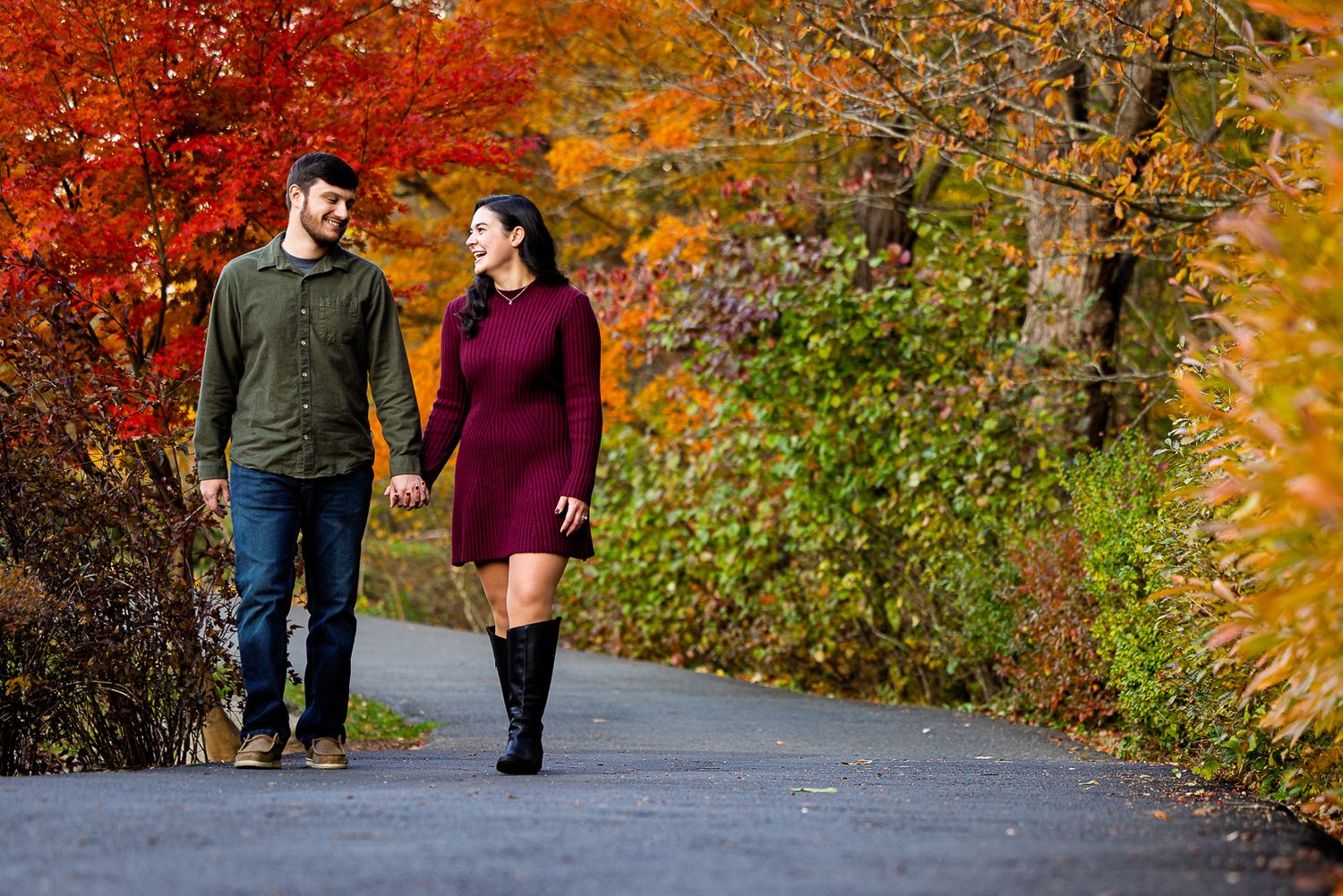 Couple holding hands walking on a path surrounded by vibrant autumn foliage in red, orange, and green.