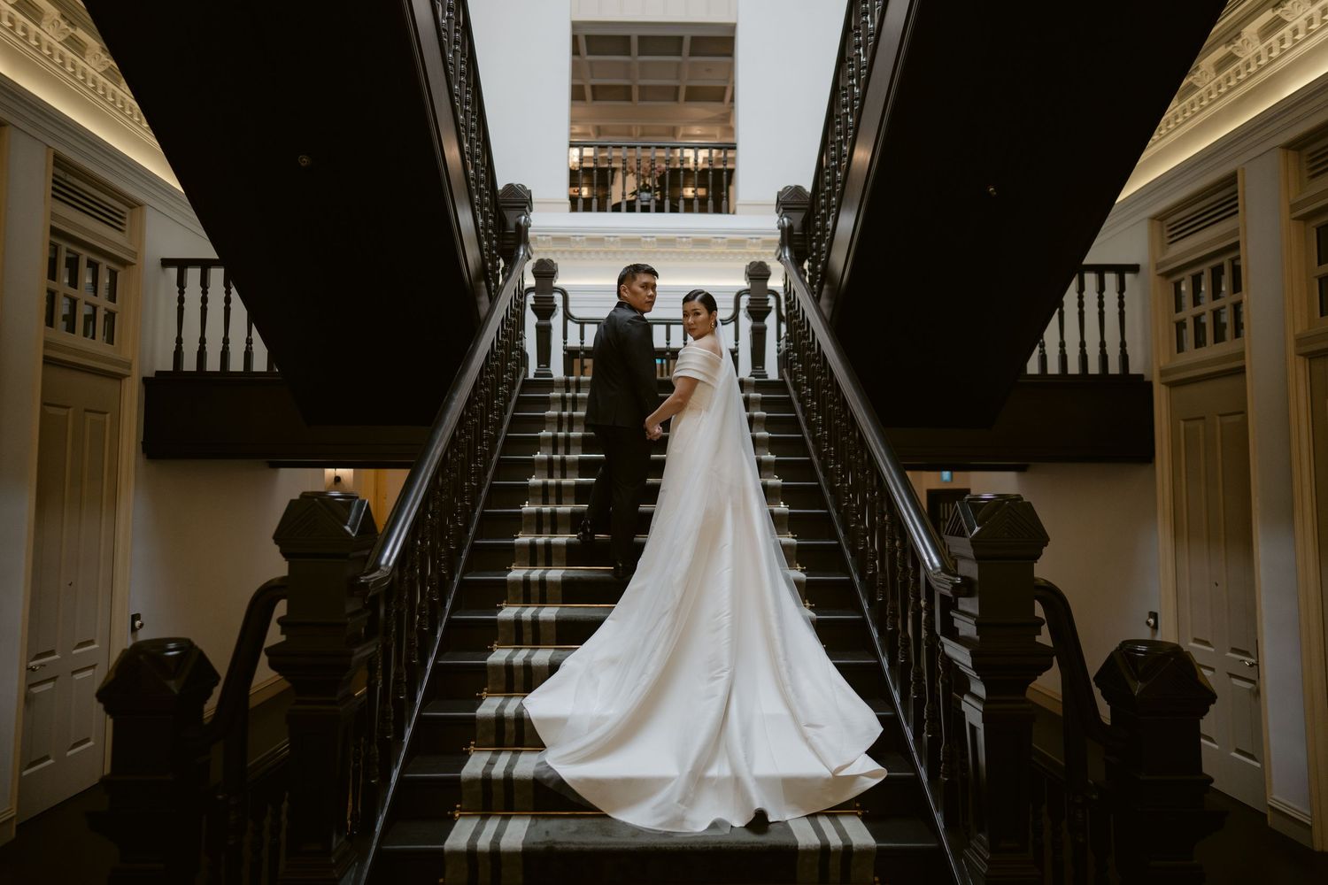 Bride and groom pose elegantly on grand staircase, bride in flowing white gown with long train in ornate venue.