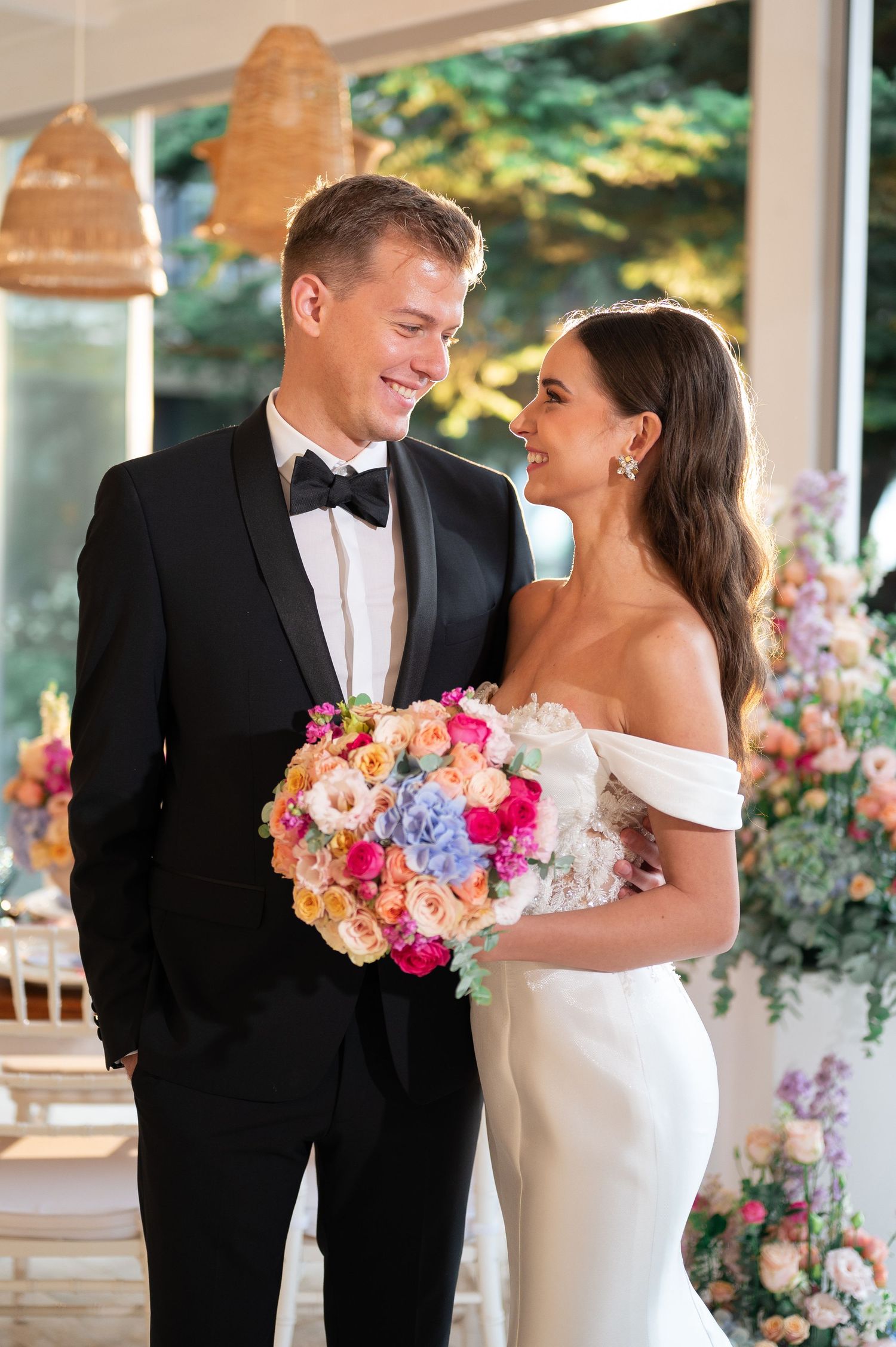 Smiling couple in elegant wedding attire with vibrant colorful bouquet in bright venue.