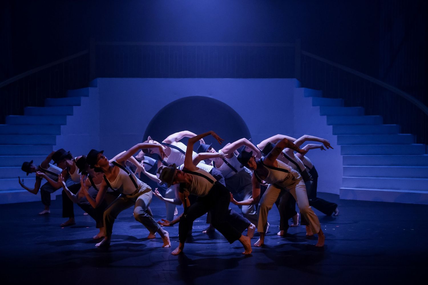 A group of dancers dressed in white tank tops, neutral-colored pants, black suspenders, and fedoras leaning forward with both arms bent.
