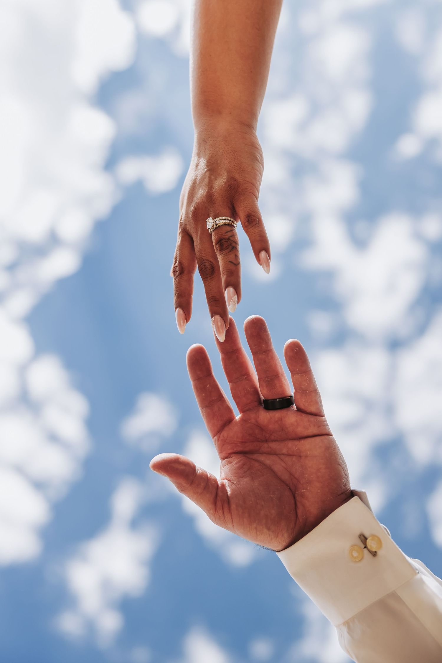 Two hands reaching toward each other against a blue sky, both wearing wedding rings, symbolizing marriage and unity.