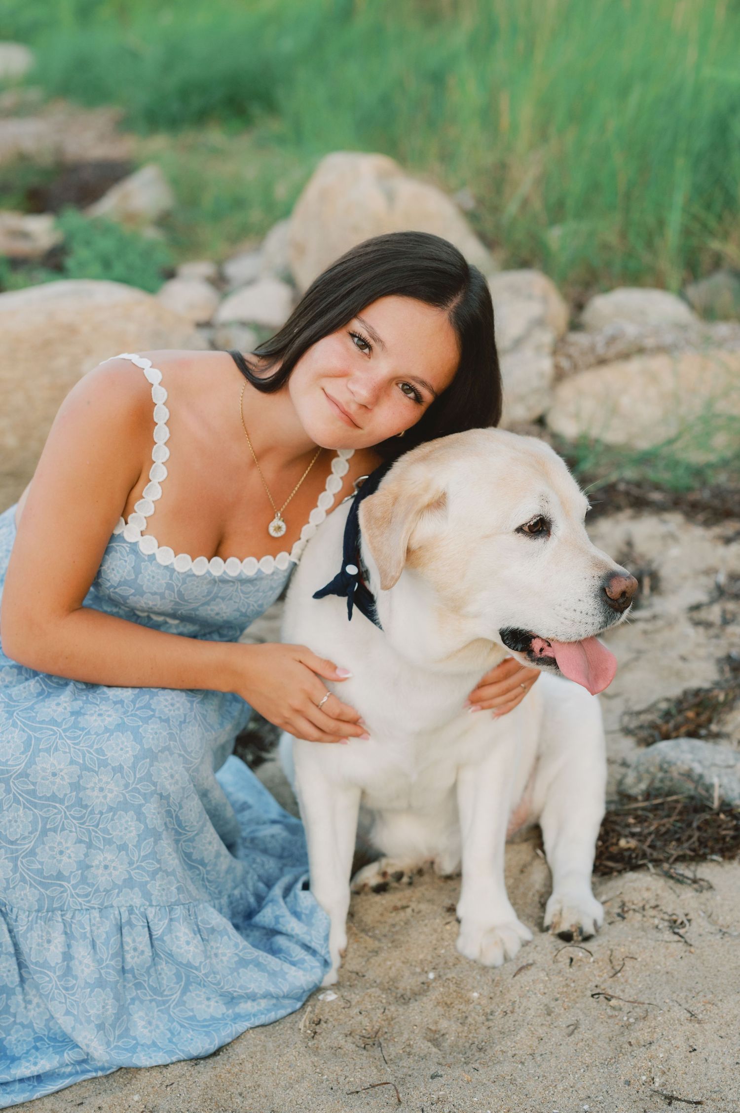 Young woman in blue floral dress smiling and hugging a yellow Labrador dog outdoors near rocks.