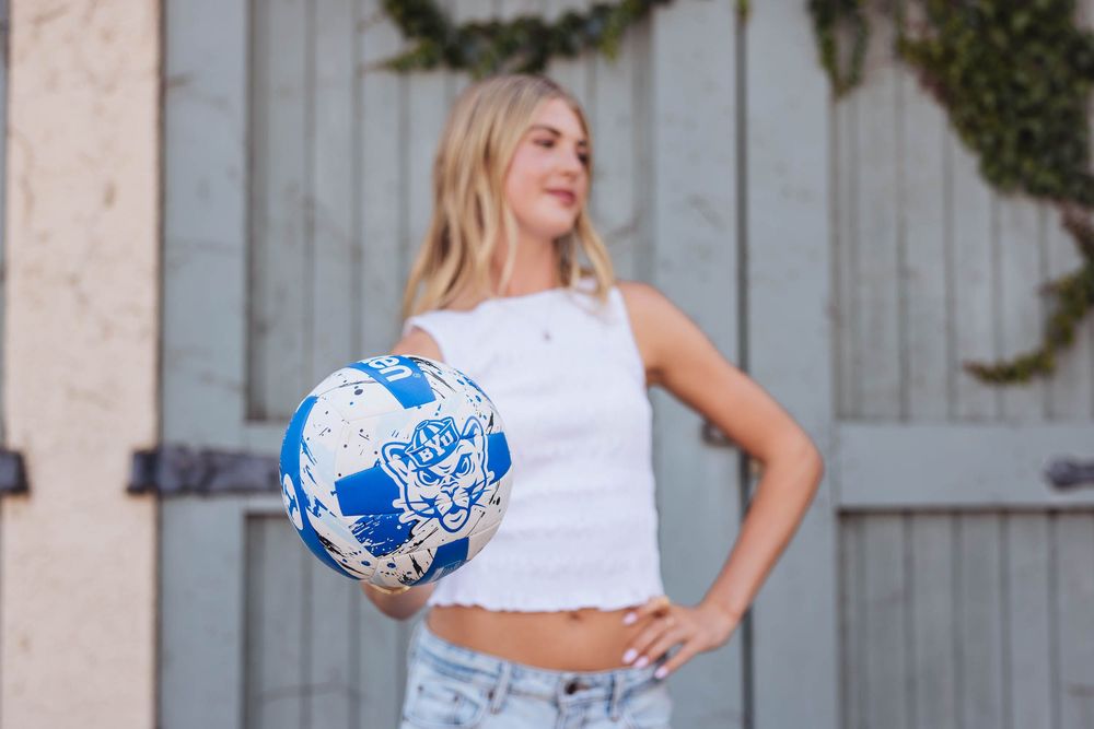 Blonde woman in white crop top holding a blue and white soccer ball outdoors near a rustic door.