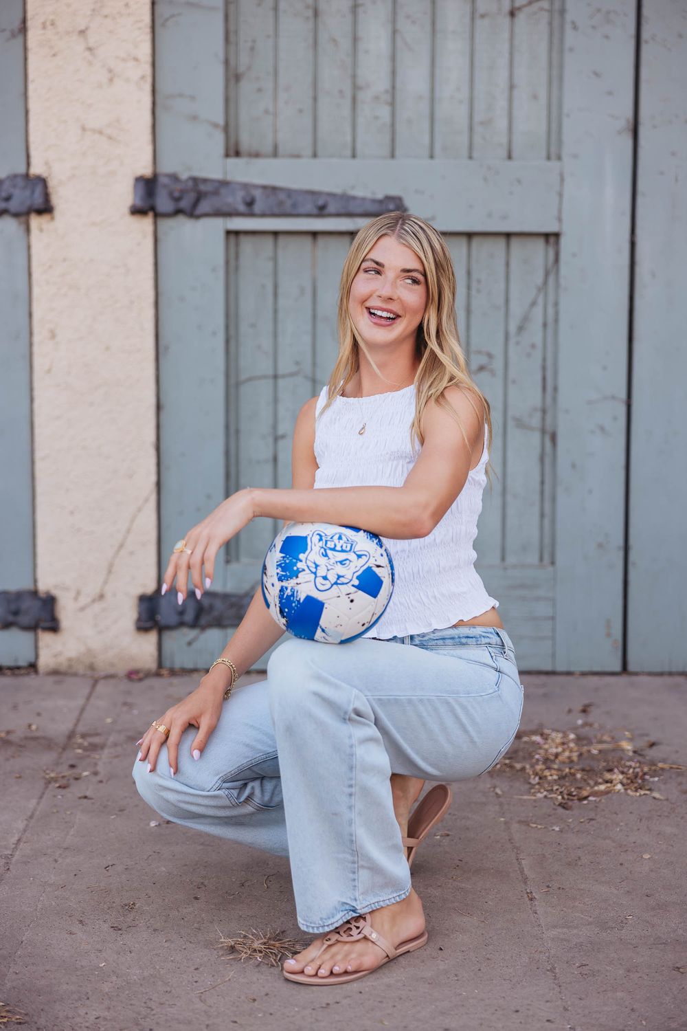 Smiling blonde woman in white tank top and jeans sitting outdoors holding a blue and white soccer ball.