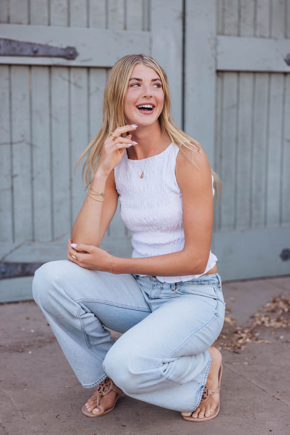 Smiling blonde woman crouching in light wash jeans and white sleeveless top with sandals outdoors.