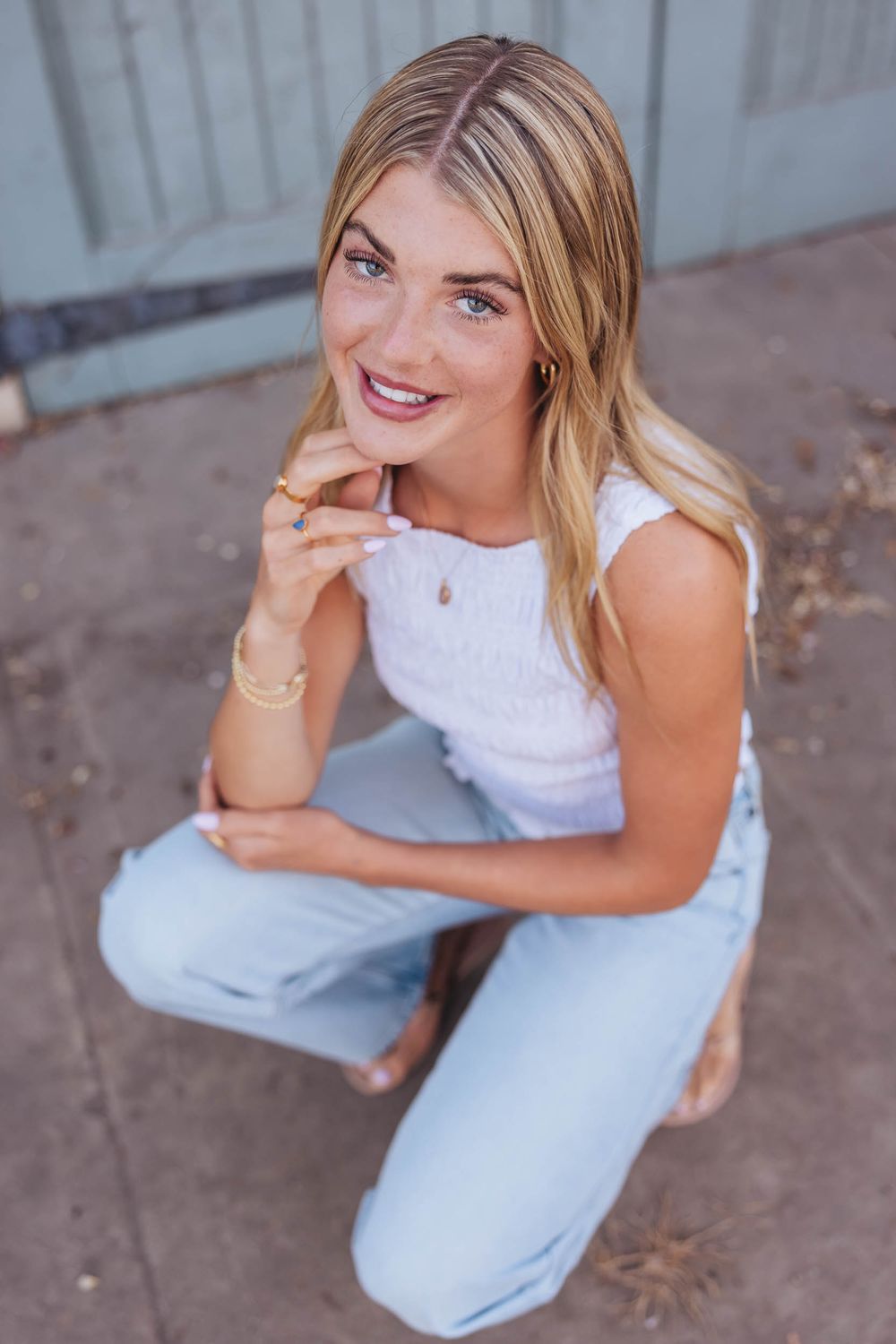 Smiling blonde woman crouching outdoors in white top and light jeans with gold jewelry.