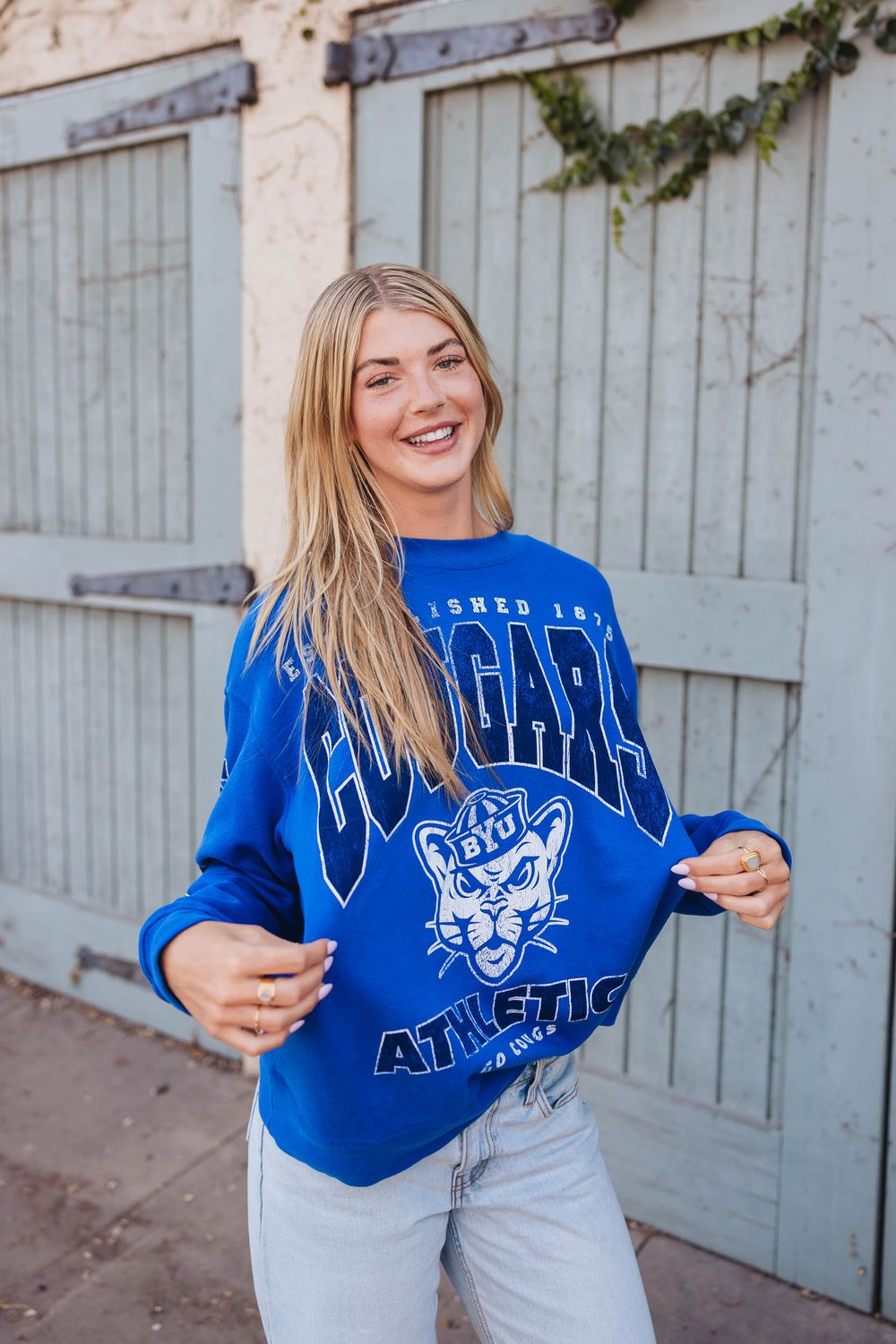 Woman smiling in a blue BYU Cougars Athletics crewneck sweatshirt, paired with light wash jeans.