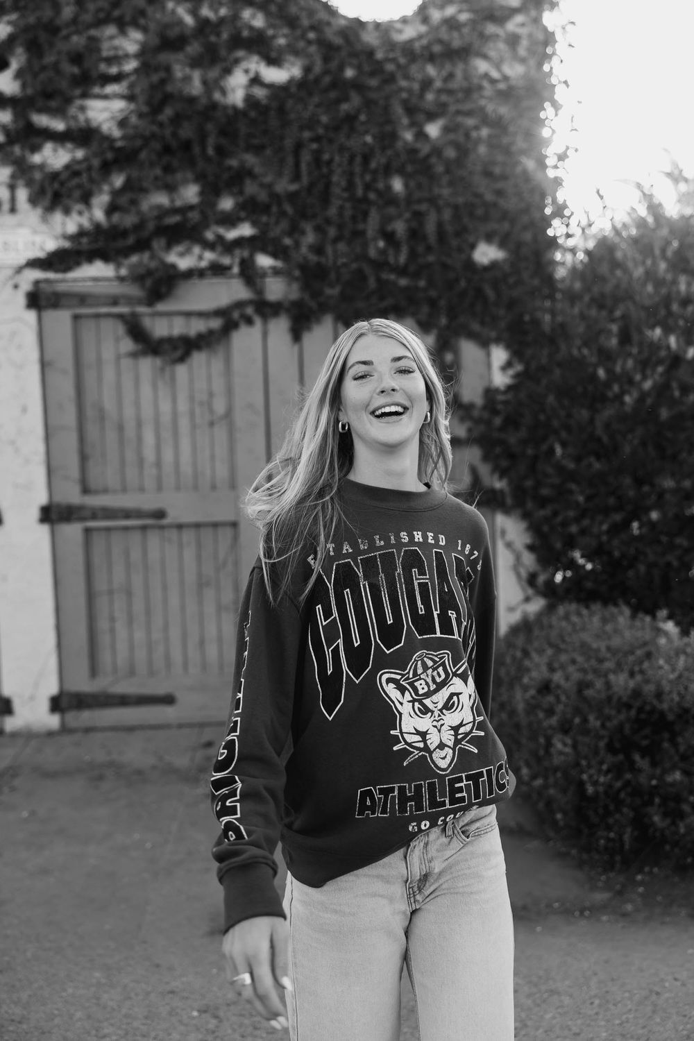 Young woman laughing outdoors in black and white, wearing a Cougars Athletics sweatshirt and jeans.