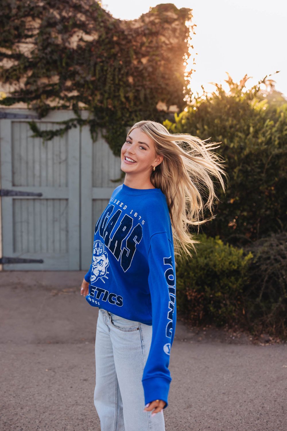 Smiling blonde woman posing outdoors in a blue Cougars Athletics sweatshirt with hair blowing in wind.