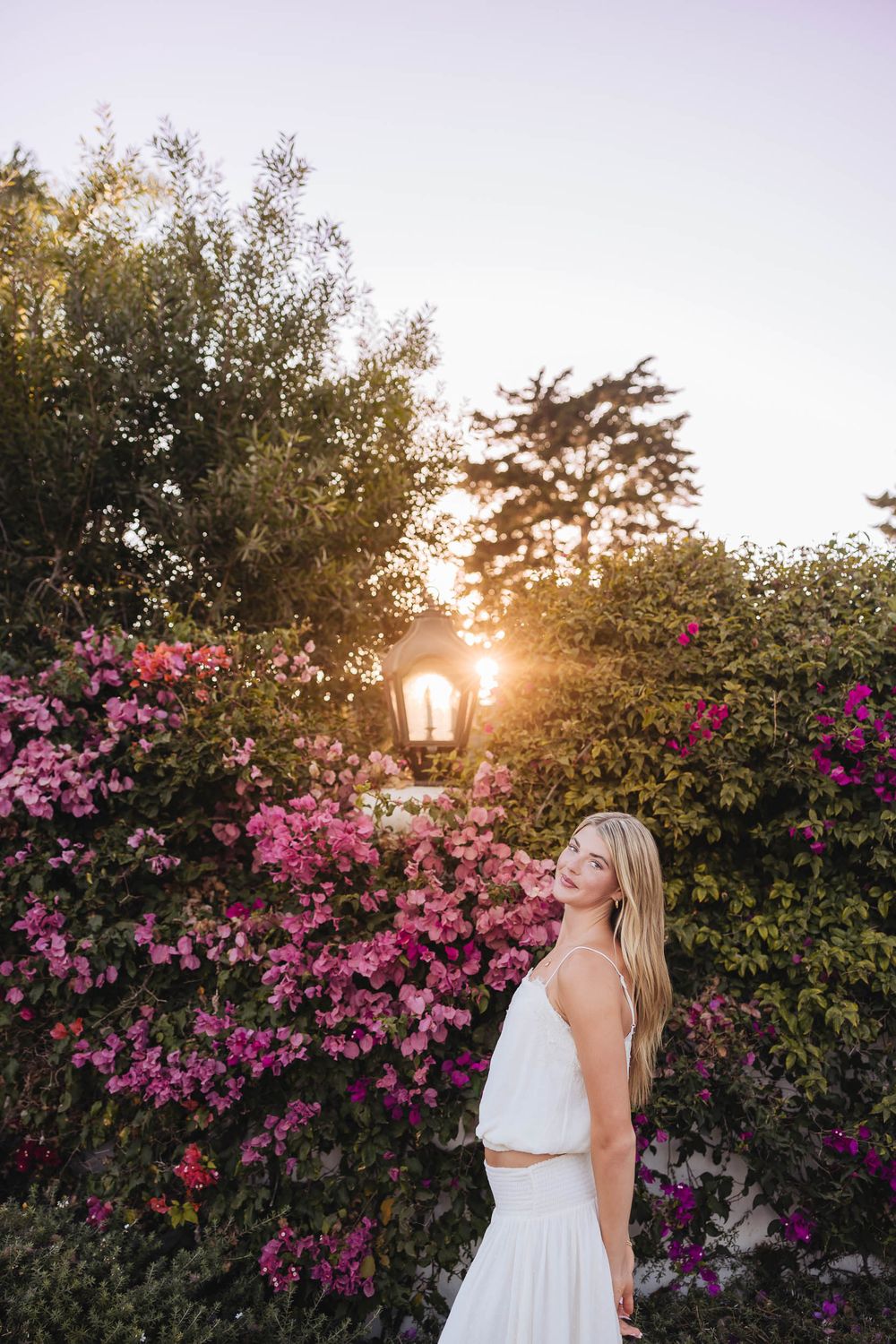 Blonde woman in white outfit smiling among pink bougainvillea flowers at golden sunset.