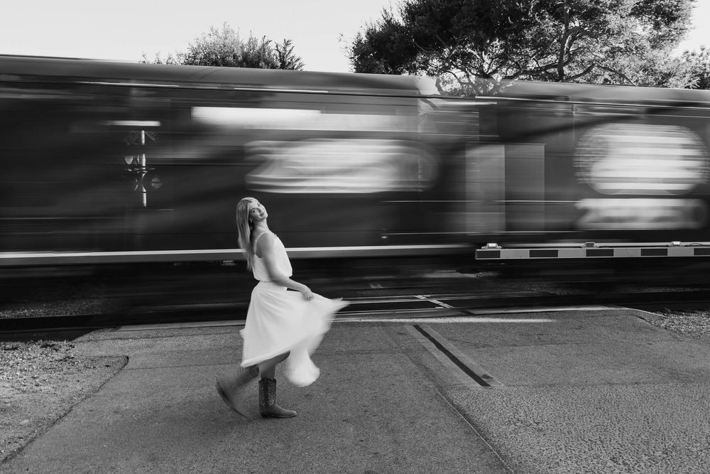 Black and white photo of woman in white dress and cowboy boots crossing railroad tracks with passing train.
