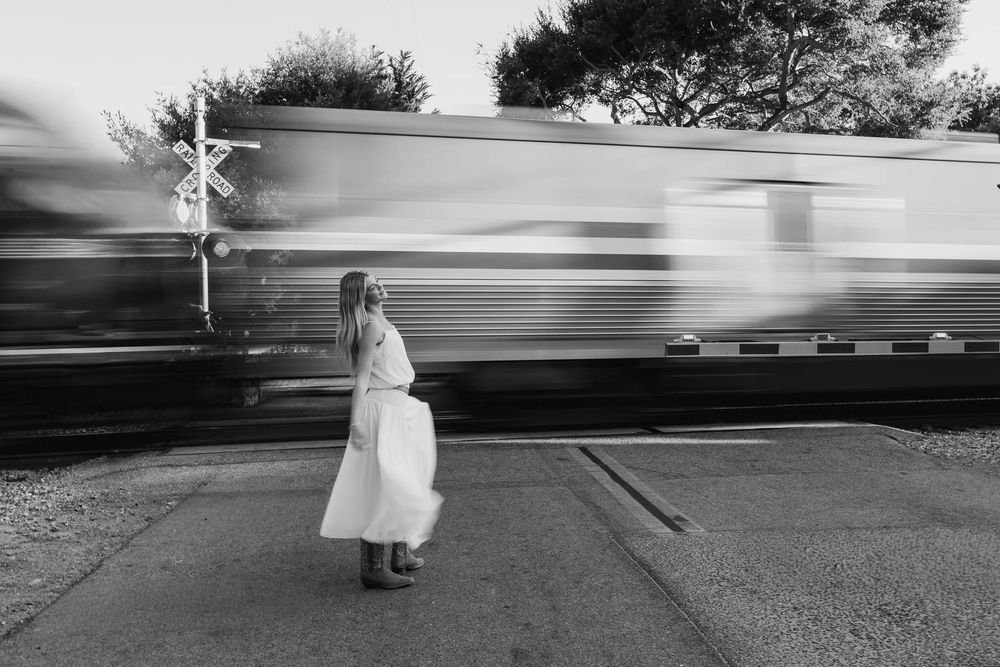 Black and white photo of woman in white dress standing at railroad crossing as train passes.