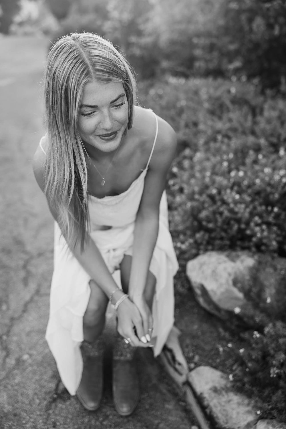 Blonde woman in white dress and boots sitting outdoors smiling in black and white photo.