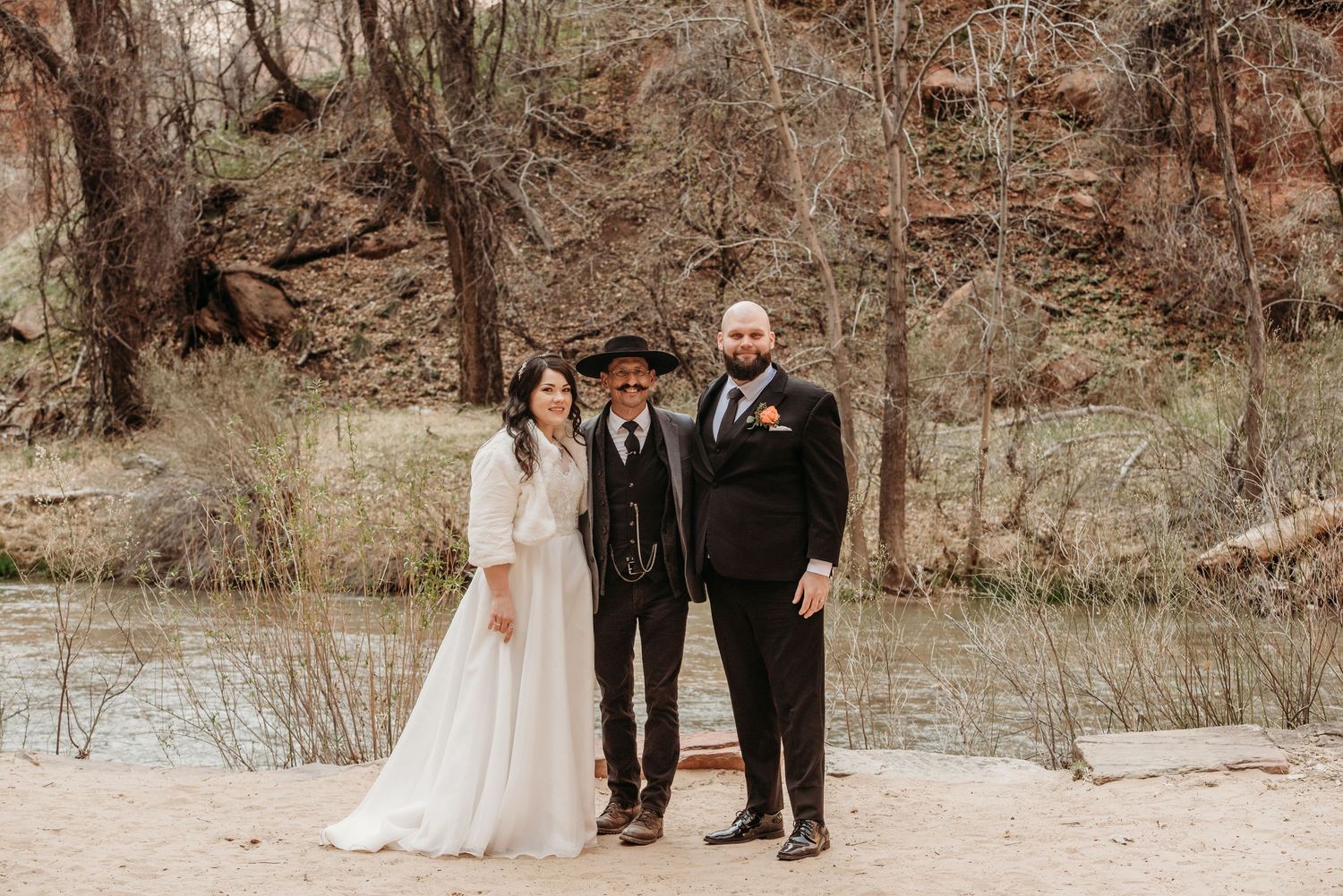 Couple in wedding attire with officiant outdoors near a river in a desert canyon setting.