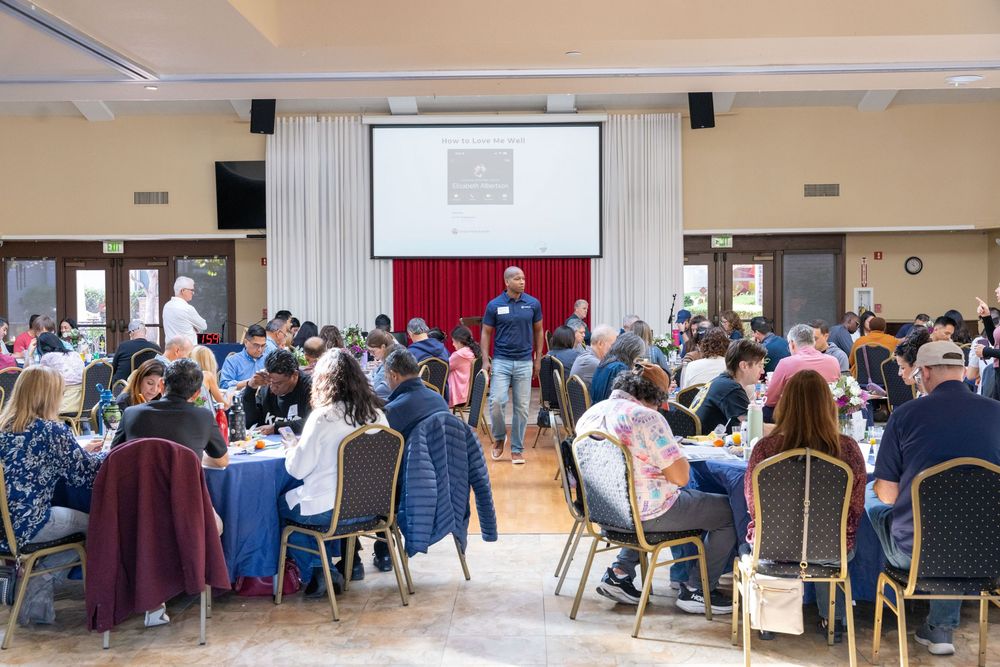Speaker presenting to a large audience seated at round tables in a banquet hall with a projection screen.