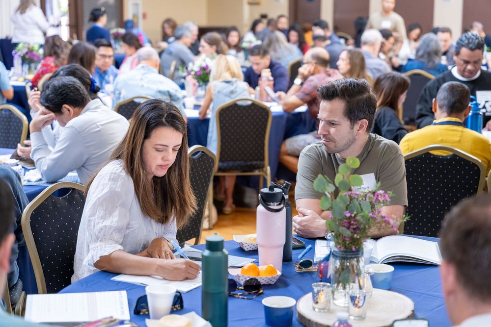 Professionals networking at a conference banquet, seated at round blue-clothed tables in a large event hall.