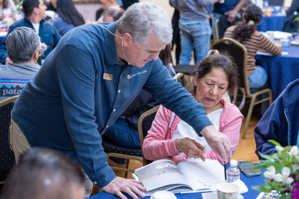 Man in blue shirt assists elderly woman reviewing documents at a busy community event gathering.