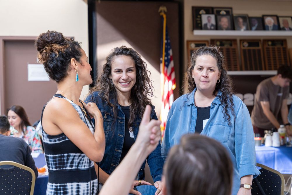 Three women smiling and conversing at an indoor gathering with an American flag and bookshelf in background.
