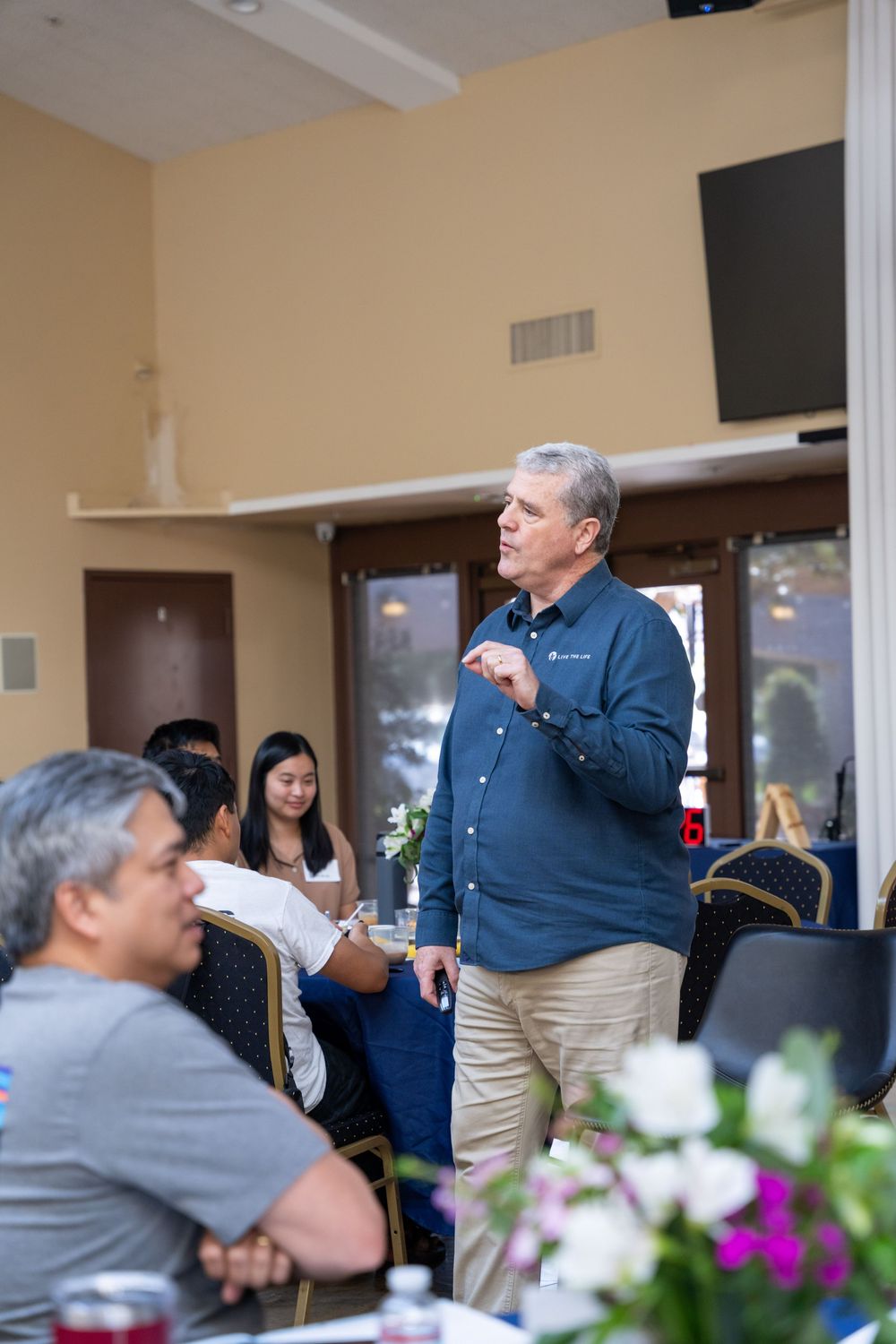 Man in blue shirt speaking to a group of people seated at tables in a banquet room.