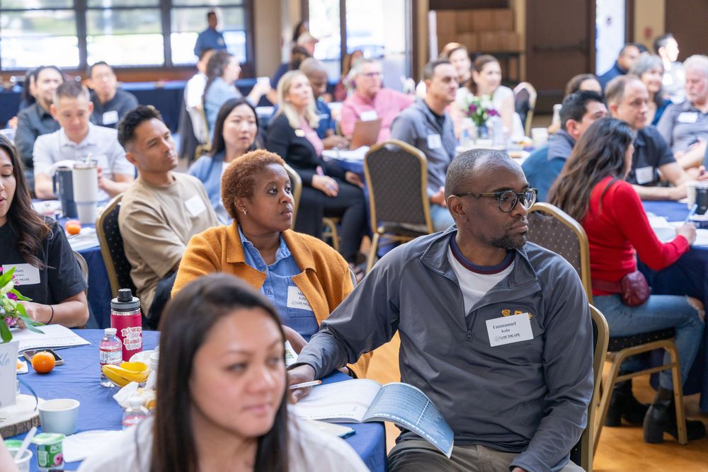 Diverse group of adults seated at round tables attending a conference or seminar in a large banquet hall.