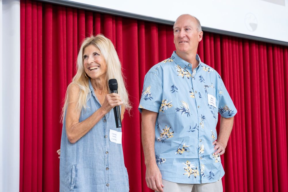 A woman with long blonde hair holds a microphone beside a man in a floral shirt, both standing before a red curtain.