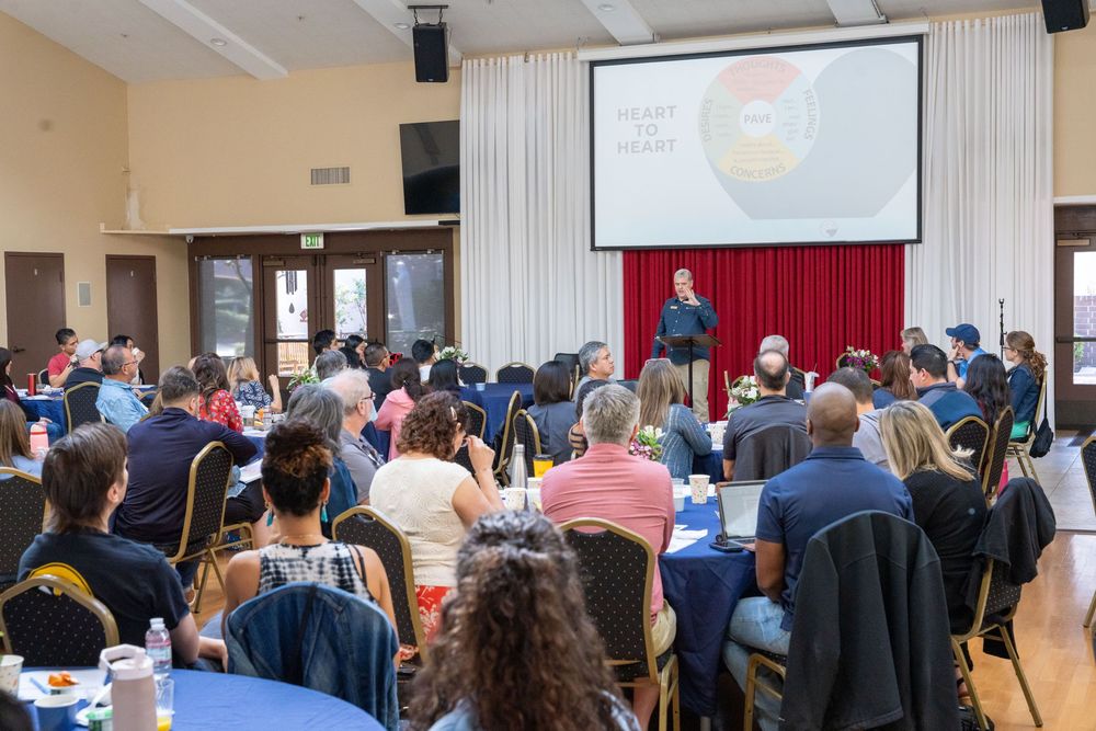 Audience seated at round tables listens to a speaker presenting on stage with a projected screen at an indoor event.