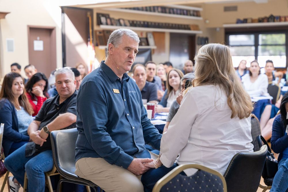 People engaged in conversation at an indoor community meeting or conference event with attendees seated in chairs.