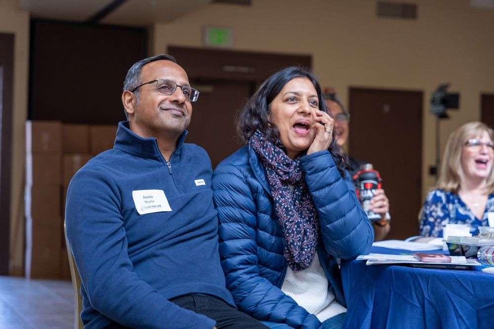 Two smiling people wearing name tags sit together at an indoor event, laughing and engaged in the presentation.