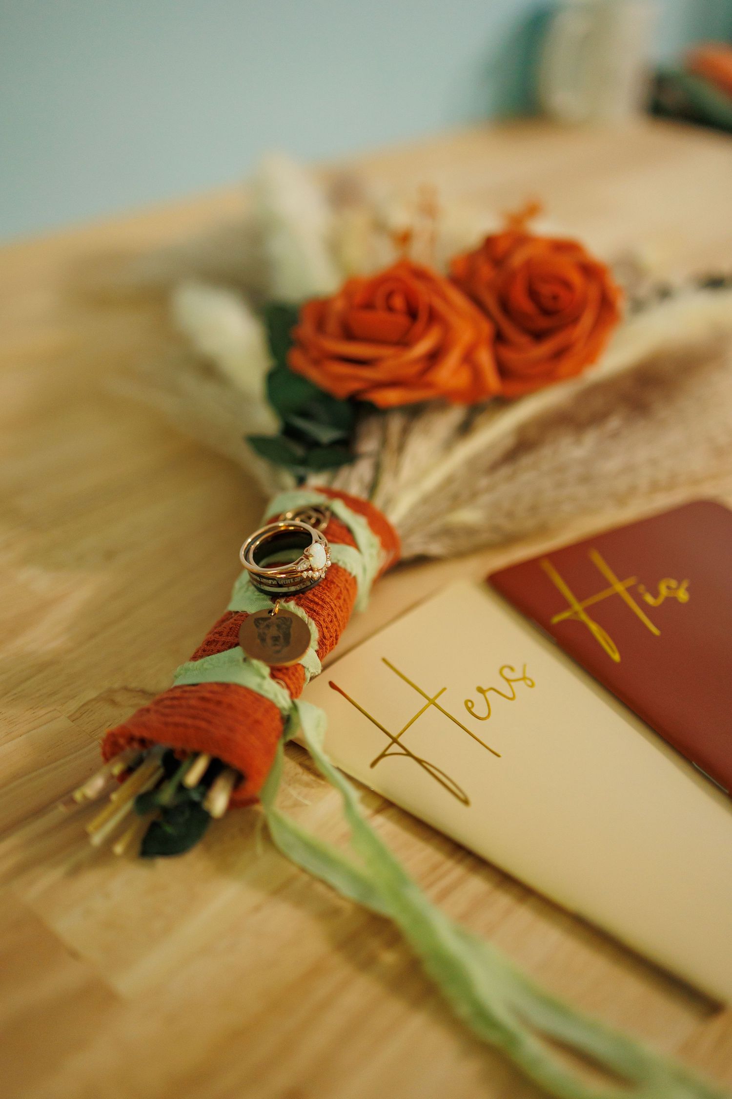 Wedding rings rest on a boho floral arrangement with burnt orange roses beside His and Hers vow books on a wooden table.