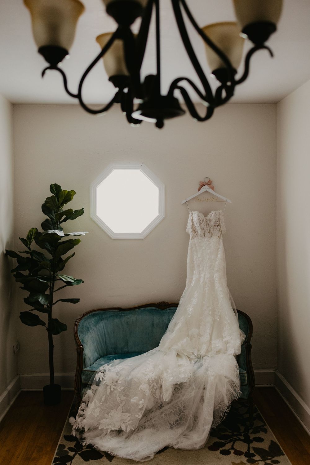 Elegant lace wedding dress hanging from a chandelier over a teal vintage settee beside a fiddle leaf fig tree.