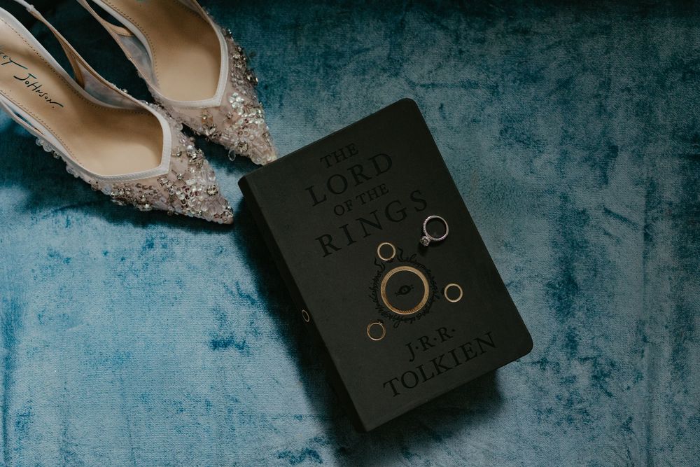 Wedding rings resting on a Lord of the Rings book by J.R.R. Tolkien beside sparkly bridal heels on blue velvet.