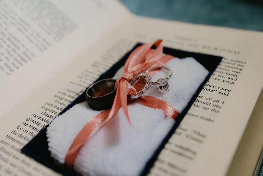 Wedding rings with a diamond engagement ring tied with a pink ribbon on a white pillow placed on an open book.