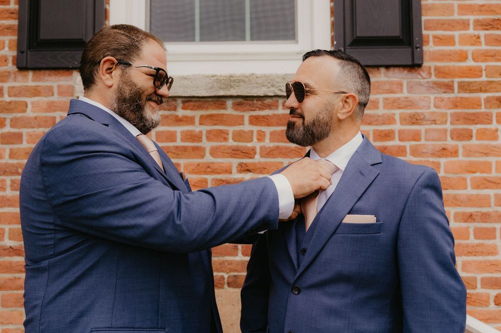 Two men in blue suits adjusting a tie outdoors in front of a brick wall.