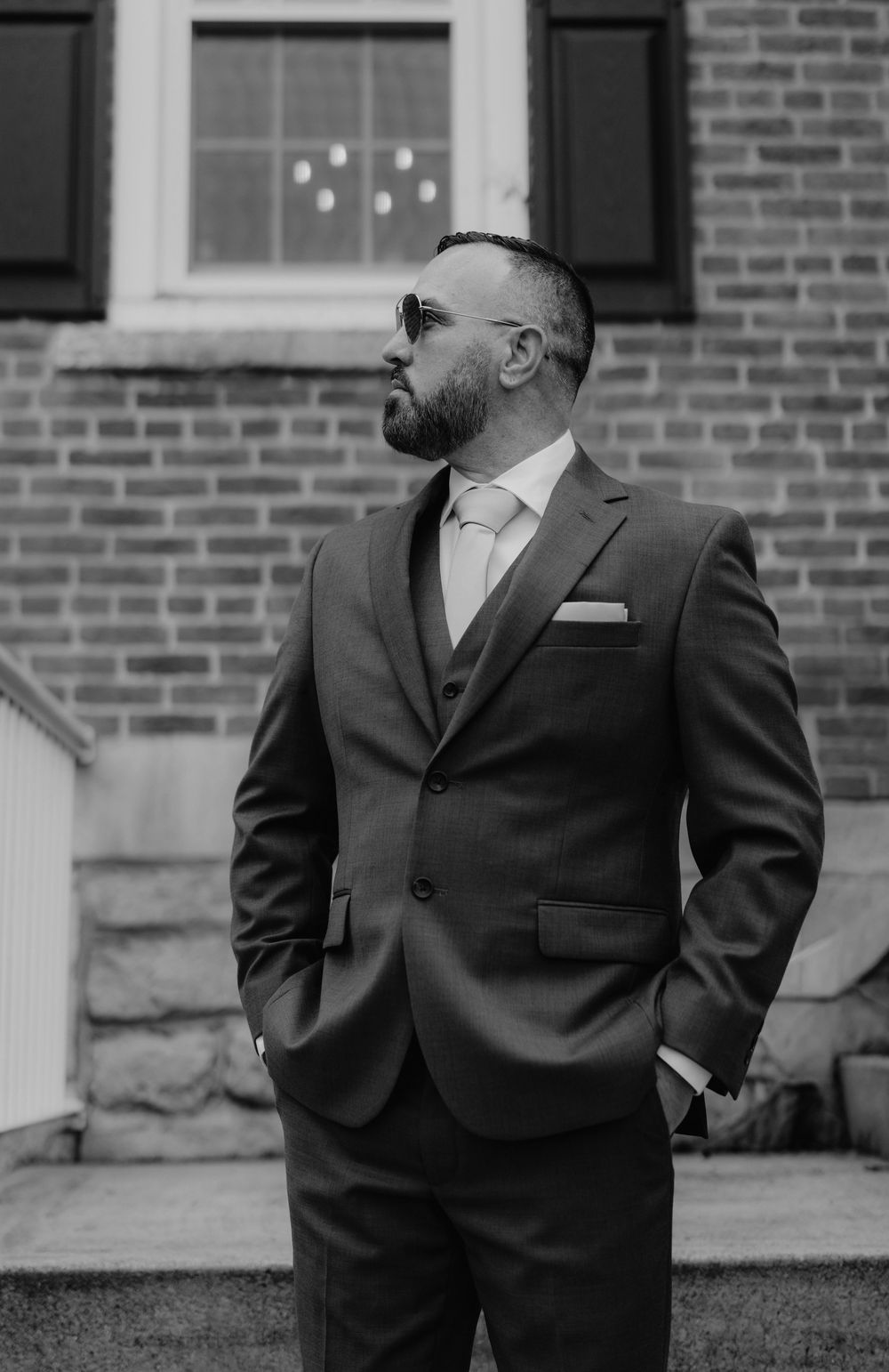Black and white photo of a well-dressed man in a suit and sunglasses standing confidently in front of a brick building.