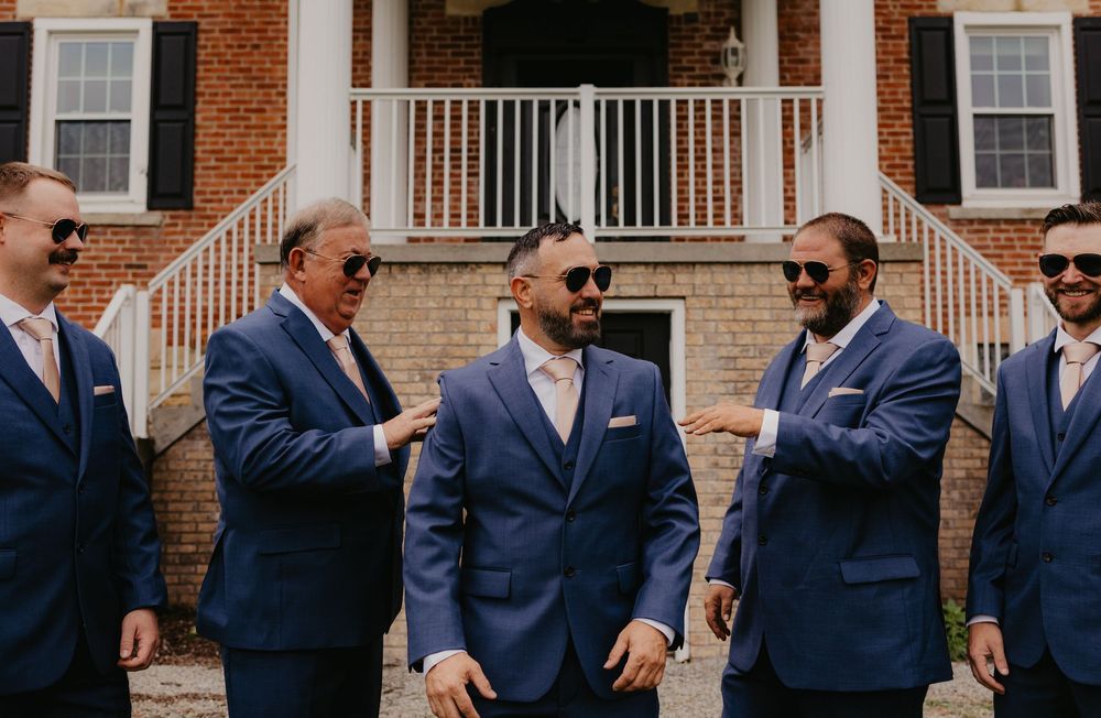Groomsmen in navy blue suits and sunglasses laughing together outside a brick building at a wedding.