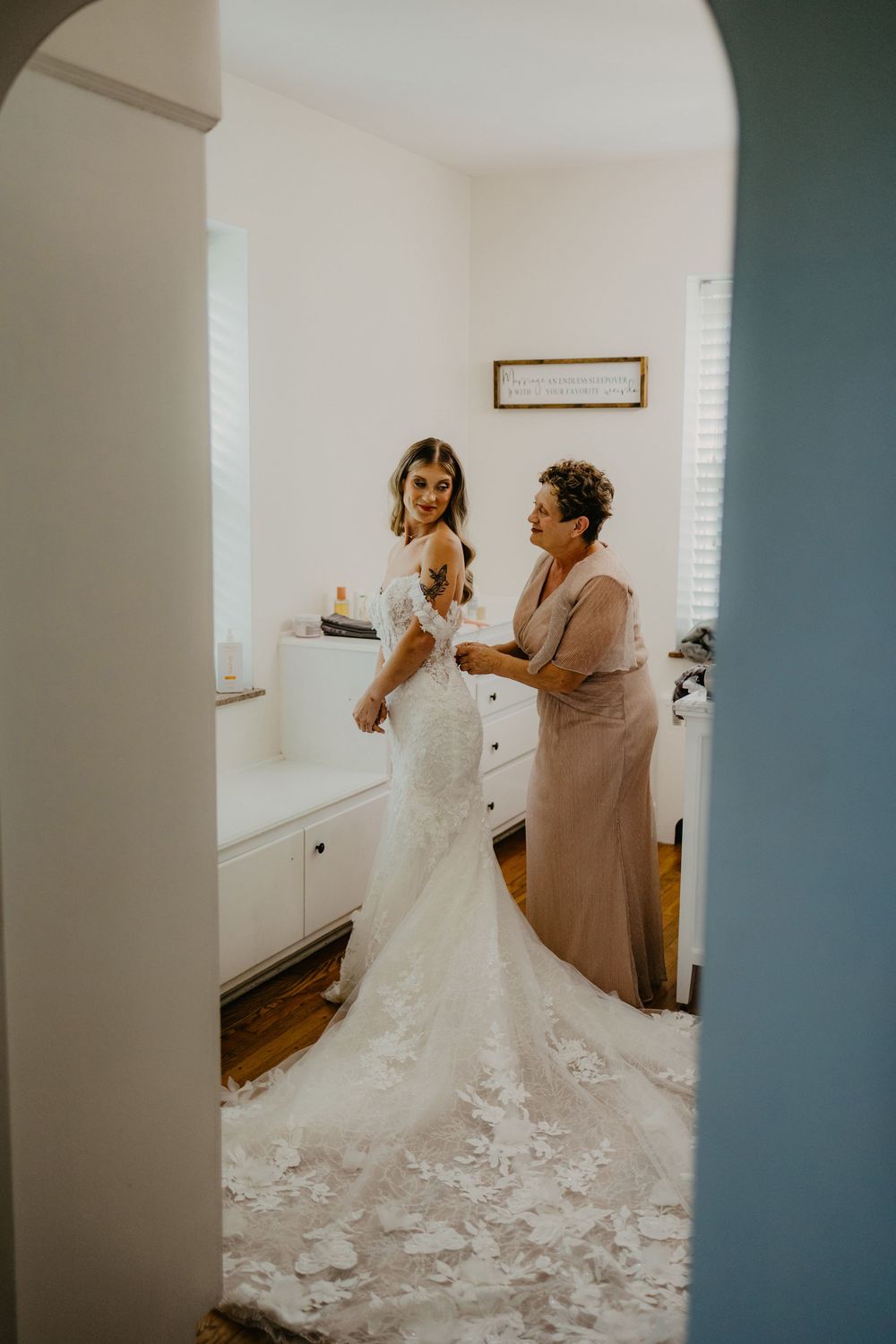 A bride in a lace mermaid wedding gown being helped by an older woman in a softly lit dressing room.
