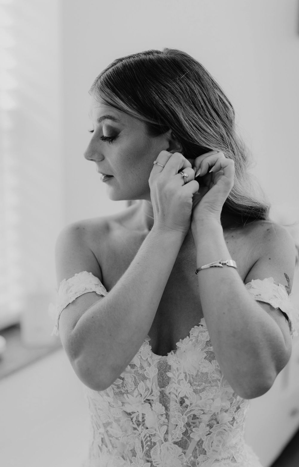 Bride in lace off-shoulder wedding gown putting on earrings, captured in elegant black and white photography.