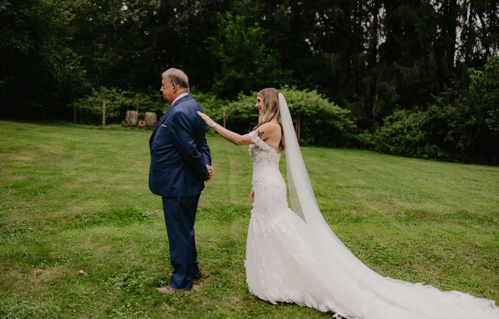 Bride in lace wedding gown and veil shares first look moment with father in blue suit on green lawn.