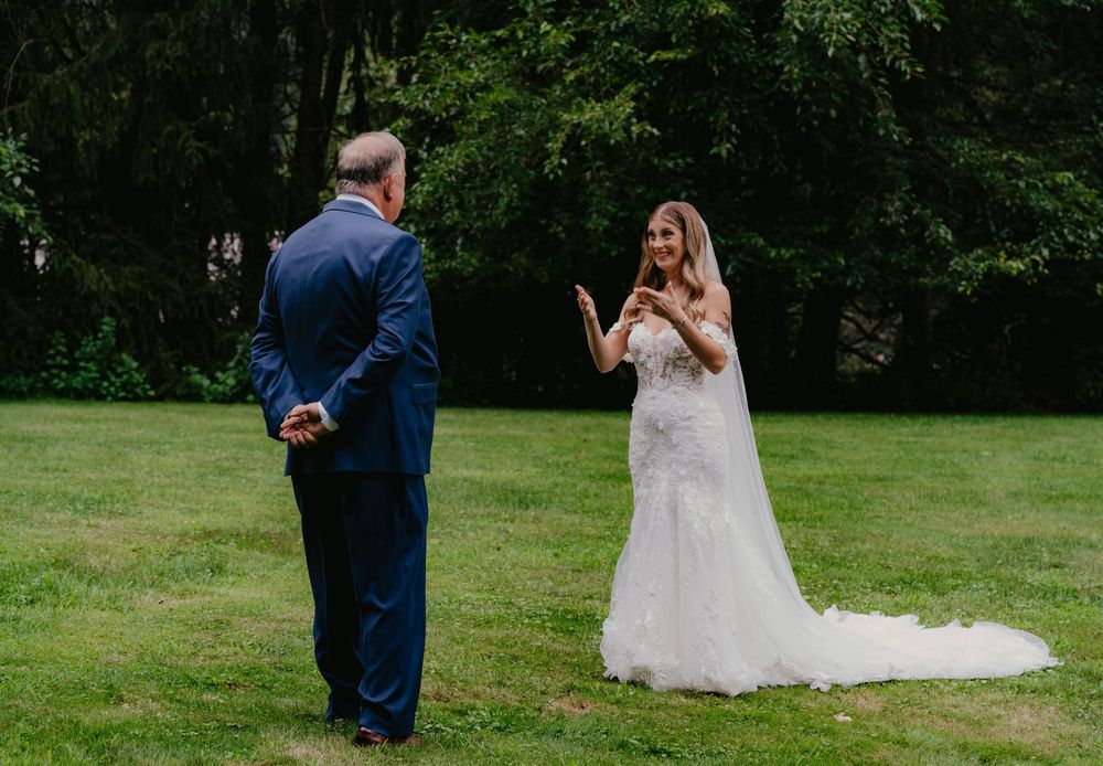 Bride in white lace gown shares emotional first look moment with suited guest on green lawn.