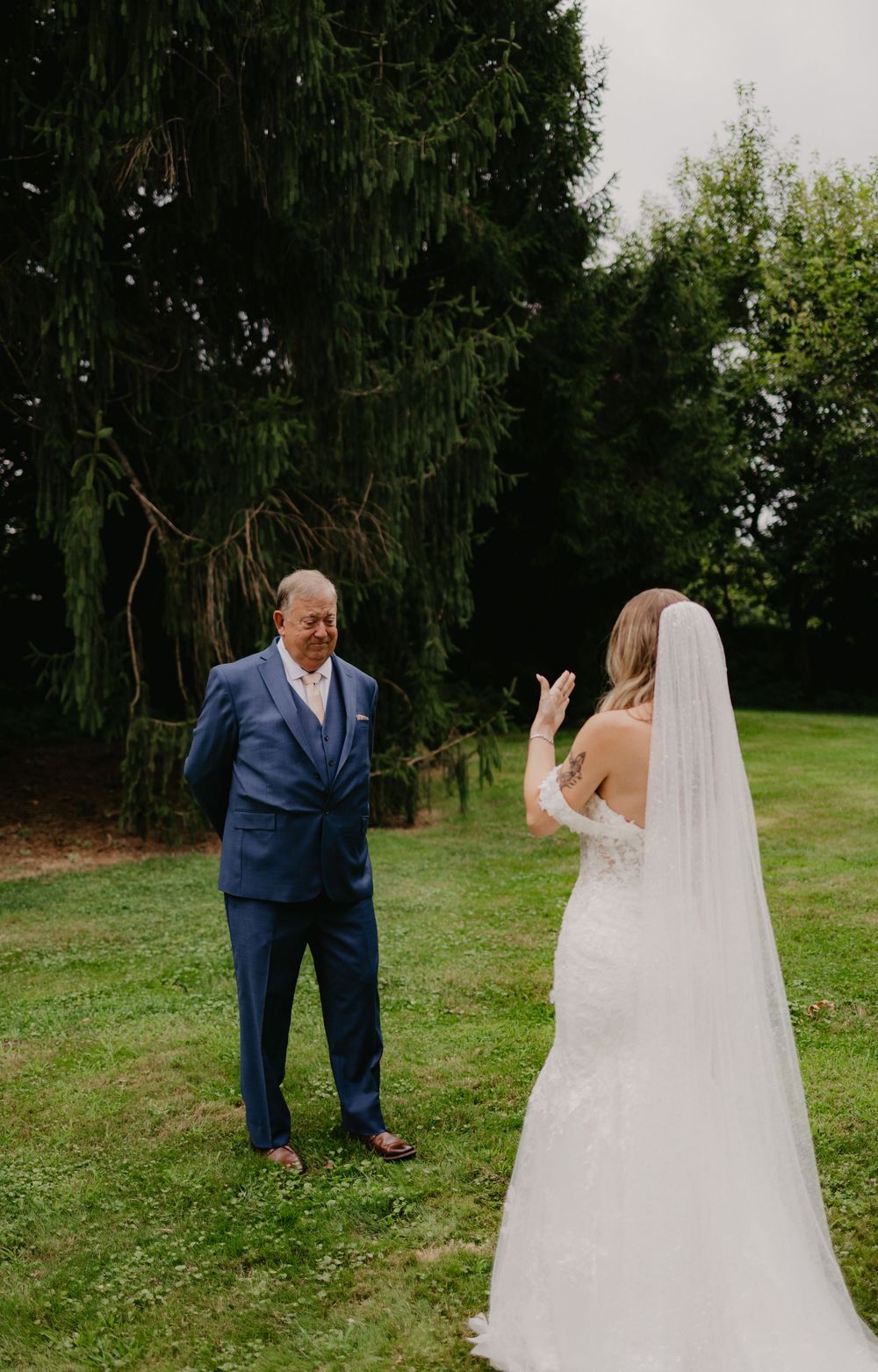 A bride in a white gown and veil shares a first look moment with a man in a blue suit on a green lawn.