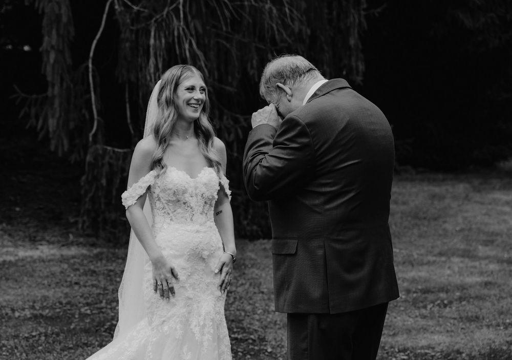 Black and white photo of a bride in lace gown sharing emotional first look moment with her father wiping tears at outdoor wedding.