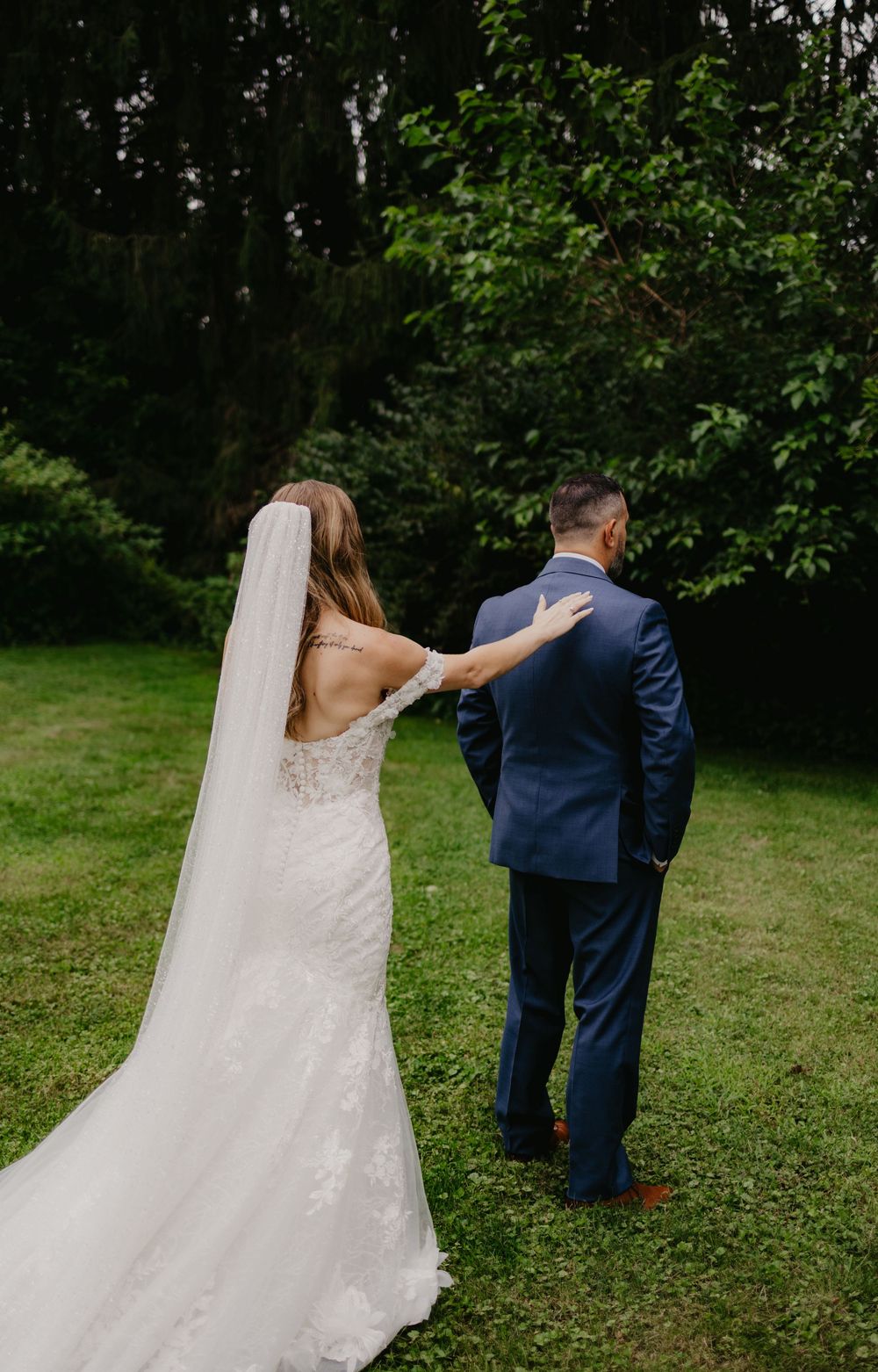 Bride in lace gown with veil touches groom in navy suit during outdoor first look on green lawn.