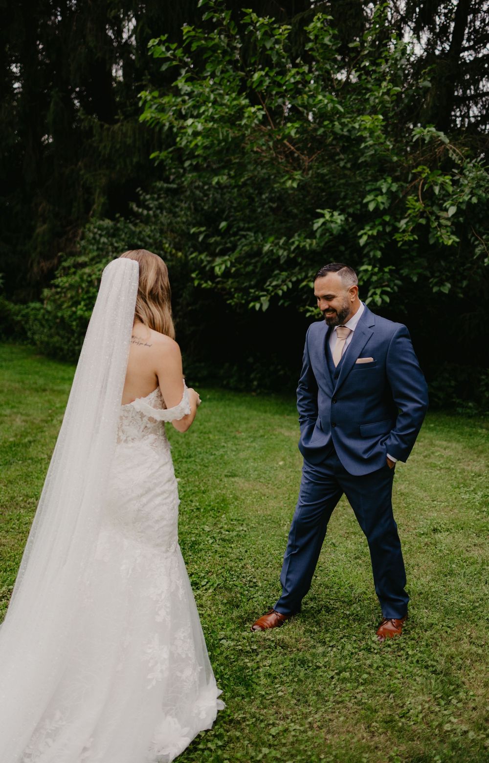 Bride in lace gown and veil shares first look moment with groom in navy suit on green lawn.