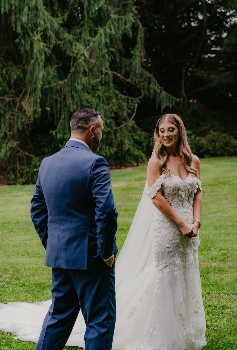 A bride in a lace off-shoulder gown smiles at her groom in a blue suit during an outdoor first look ceremony.