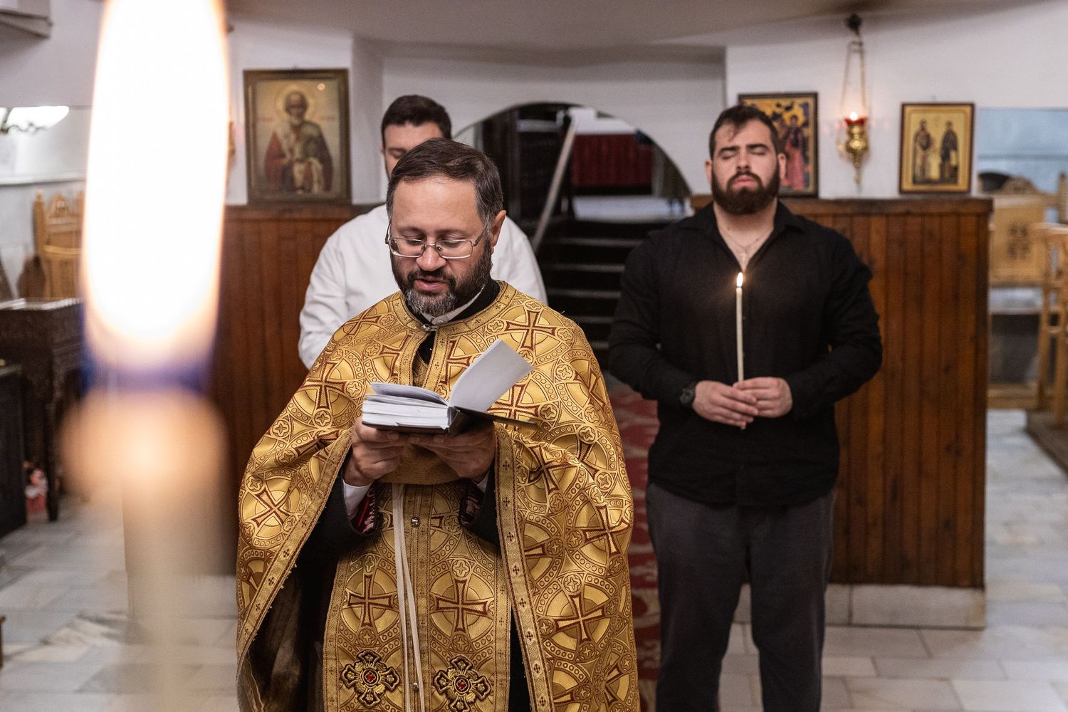 Orthodox priest in golden vestments reads scripture during a religious ceremony in a decorated church.