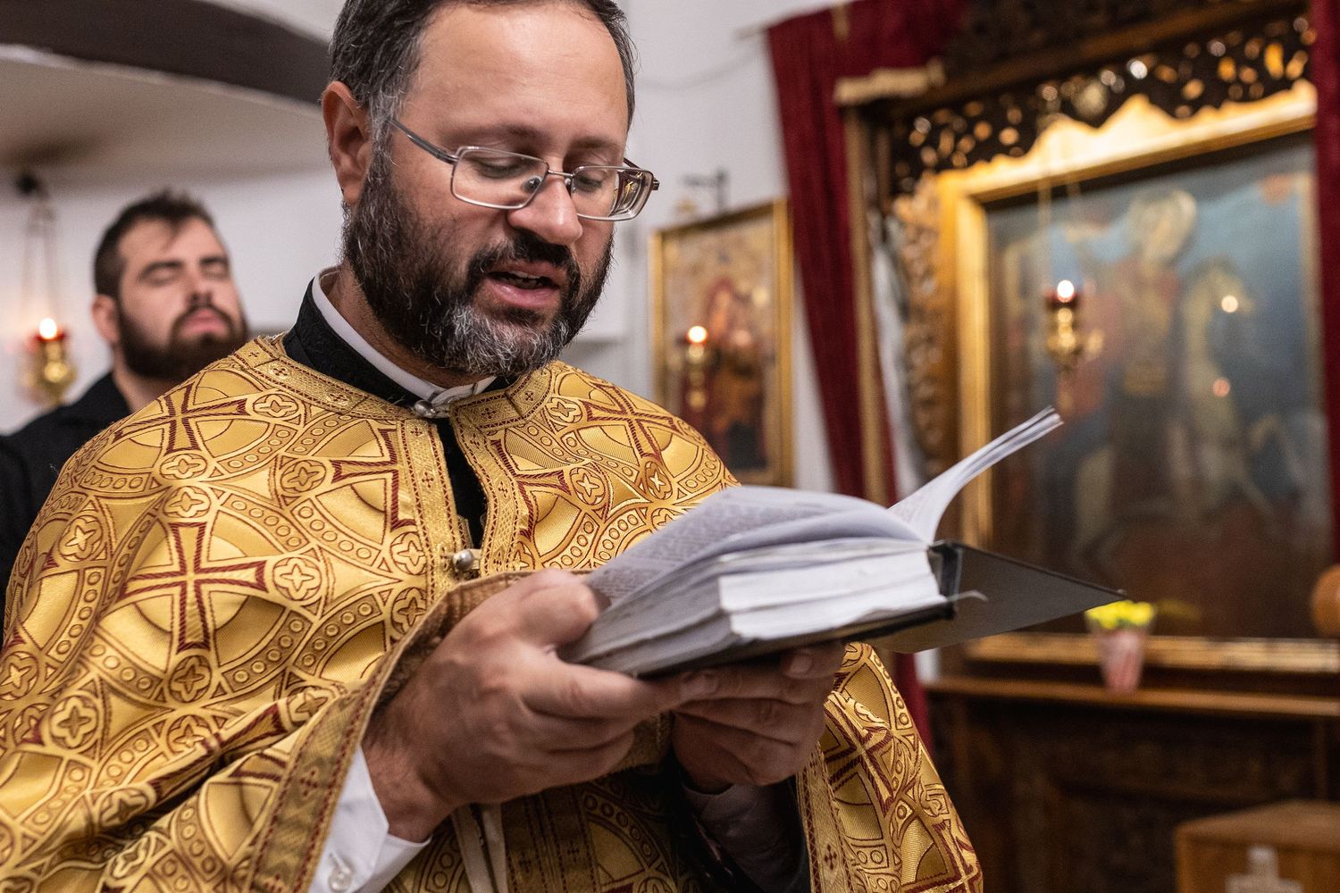 Orthodox priest in golden vestments reads from a liturgical book during a religious ceremony in a decorated church.