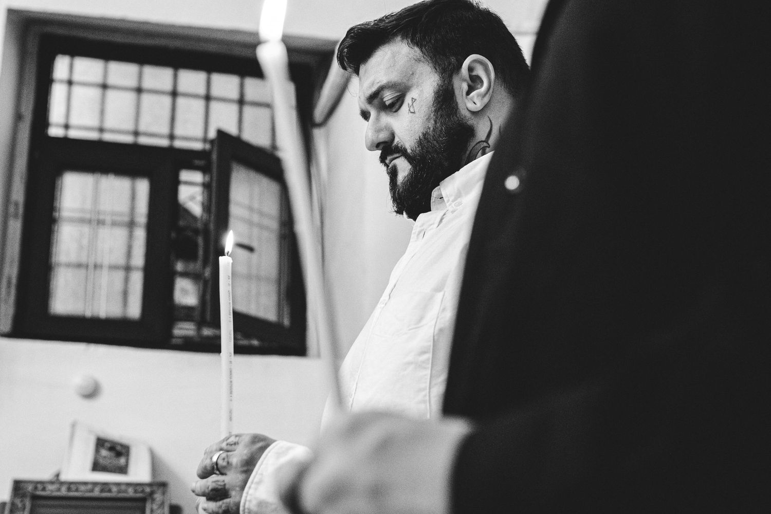 Black and white photo of a bearded man in a white shirt looking down thoughtfully near a window.